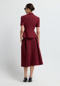 Donna che indossa un blazer attillato bordeaux a maniche corte e una gonna midi abbinata, tacchi neri, orecchini d'oro e occhiali da sole neri, in piedi di lato.