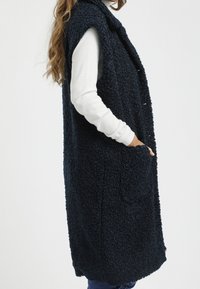 Granatowa, bezrękawnik z futrzanym wykończeniem i kołnierzem, z dwoma bocznymi kieszeniami; noszony na długim, białym koszuli.