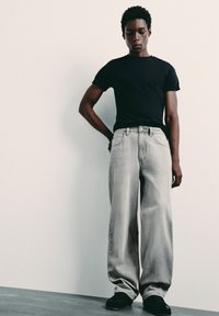 Personne debout contre un mur uni, portant un t-shirt noir, un jean large gris clair et des chaussures noires, regardant vers le bas avec une main derrière le dos.