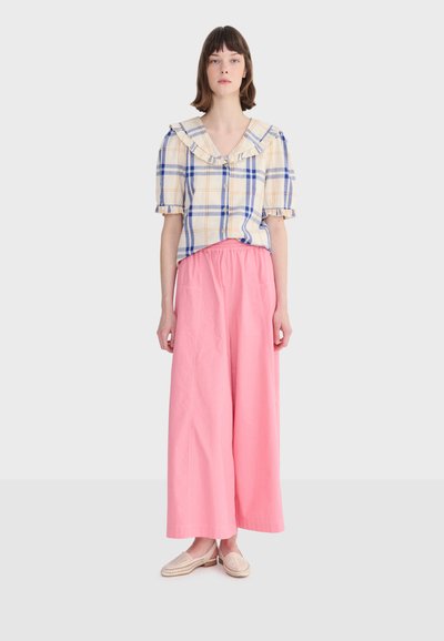 Jeune femme portant un chemisier à carreaux bleu et jaune avec col à volants, pantalon large rose, et chaussures beige à enfiler, debout droite devant un fond blanc.