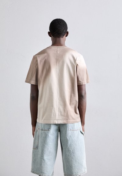 T-shirt à manches courtes, dégradé beige avec un col rond, fabriqué en coton doux. Porté avec un short en denim clair comportant de grandes poches.