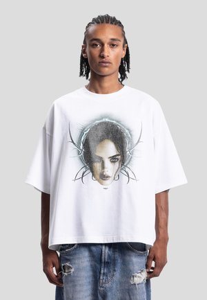 Mann med fletter som har på seg en oversized hvit T-skjorte med et mørkt kunstnerisk ansikt med abstrakte sirkulære linjer, kombinert med revne blå jeans.