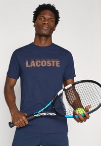Μπλε ναυτικό t-shirt με το λογότυπο "LACOSTE" σε ανάγλυφο καφέ σχέδιο. Το μοντέλο κρατάει ένα ρακέτα τένις και μια πράσινη μπάλα τένις.