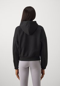 Čierna mikina s kapucňou a skráteným strihom, s elastickým lemom a manžetami. Hladká textúra látky a uvoľnený strih. Šedé legíny sú viditeľné nižšie.