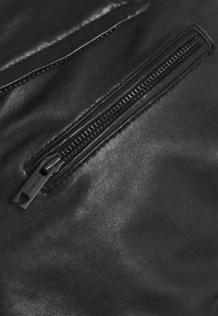 Zwart leren stof met een gladde textuur, voorzien van een zichtbare ritssluiting met metallic afwerking en een klein label aan de ritstrekker.