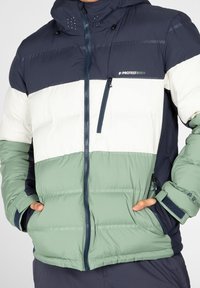 Padded jacket with a navy, white, and green color block design. Features a front zip, pockets, and a hood, made from lightweight material.