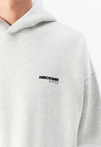 Jasnoszara bluza z kapturem z czarnym haftowanym logo "Abercrombie & Fitch" na klatce piersiowej, noszona przez osobę na białym tle.