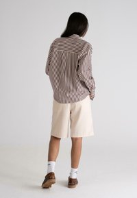 Stribet brun og hvid skjorte med knaplukning, parret med beige shorts. Modellen bærer hvide sokker og brune sko. Neutral baggrund.