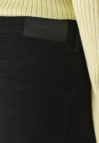 Crne traperice s teksturiranim materijalom, džepovima s pločicama i kožnom etiketom s natpisom "BLANCHE." Outfit je upotpunjen žutom rebrastom duksericom.