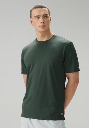 Nuori mies, jolla on päällään tummanvihreä lyhythihainen urheilupaita ja valkoiset shortsit, katsoo vasemmalleen harmaan tasaisen taustan edessä.