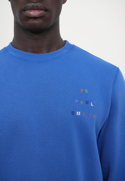Pull bleu avec un col rond et des poignets côtelés. Texte brodé multicolore remarquable sur la partie gauche de la poitrine. Matière en mélange de coton.