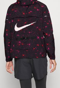 Zwarte jas met een levendig roze en rood abstract patroon, voorzien van een wit Nike-logo, elastische manchetten en een detail van een voorzak.