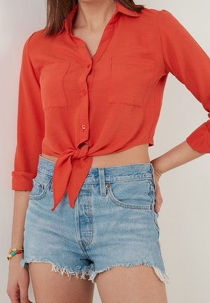 Femme portant un crop top orange à manches longues boutonné et noué, avec deux poches poitrine et un short en jean bleu clair effiloché.