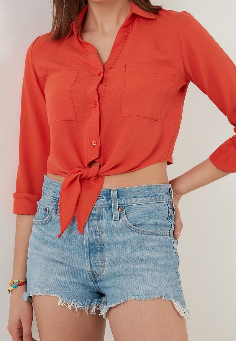 Femme portant un crop top orange à manches longues boutonné et noué, avec deux poches poitrine et un short en jean bleu clair effiloché.