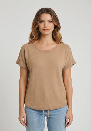 Femme aux cheveux ondulés mi-longs portant un haut en maille beige à manches courtes avec les poignets retroussés et un jean bleu, debout devant un fond uni.