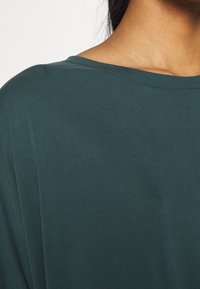 Tealfärgad bomullstopp med avslappnad passform, vid halsringning och mjuk textur som visar en del av axeln och nackområdet.