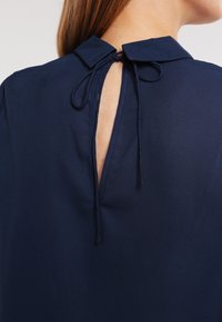 Blusa blu navy con colletto e chiusura a cravatta sul retro. Il tessuto è liscio, con un'apertura a chiave sulla nuca.