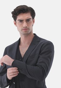 Schwarze Blazer mit Reverskragen, Ein-Knopf-Verschluss und aufgesetzter Tasche, kombiniert mit einem schwarzen V-Ausschnitt-Pullover darunter.