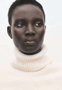 Hellebeige Rollkragenpullover mit gerippter Textur, getragen von einer Person mit kurzen, dunklen Haaren und glatter, dunkler Haut vor einem hellen Hintergrund.