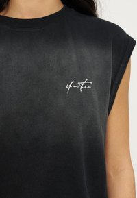 Μαύρο αμάνικο πουκάμισο με λευκό γραφικό κείμενο "You&From" στο πάνω αριστερό μέρος του στήθους, φορεμένο από άτομο με σκουρόχρωμα μαλλιά.