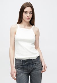 Νεαρή γυναίκα με μακριά καστανά μαλλιά, που φοράει λευκό αμάνικο τοπ και γκρι ψηλόμεσα τζιν, με φόντο ένα απλό φόντο.