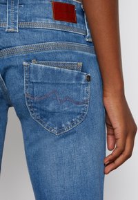 Blå denimjeans med en bakficka med en subtil vågmönstrad söm, brun läderetikett och kontrasterande sömnad. Slät textur.