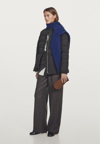 Giubbotto imbottito nero con zip, sciarpa blu oversize, pantaloni larghi marrone scuro e guanti in pelle marrone. Il modello è in piedi contro uno sfondo neutro.