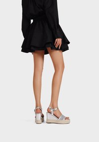 Sandali con zeppa in un upper metallizzato argentato, con dettagli di cinturini glitterati e una suola in juta intrecciata. Gonna nera con orlo arricciato sullo sfondo.