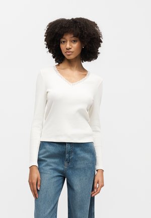 Γυναίκα με σγουρά μαλλιά που φορά λευκή μπλούζα με μακριά μανίκια και δαντελωτή διακόσμηση και μπλε τζιν με ψηλή μέση, στέκεται μπροστά σε απλό φόντο.