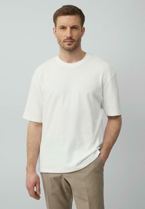 Weißes, kurzärmeliges, strukturiertes T-Shirt mit rundem Ausschnitt, kombiniert mit hellbraunen Hosen. Der Stoff weist ein dezentes geometrisches Muster auf.