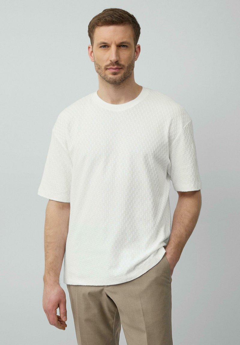 Weißes, kurzärmeliges, strukturiertes T-Shirt mit rundem Ausschnitt, kombiniert mit hellbraunen Hosen. Der Stoff weist ein dezentes geometrisches Muster auf.