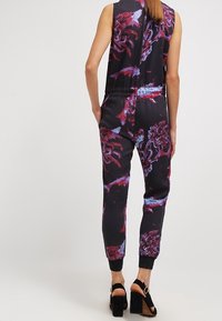 Svart jumpsuit med blommönster i nyanser av lila och röd, med en figursydd midja, elastiska ärmslut och öppen ryggdesign.