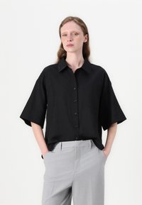 Camisa preta de manga curta com um design de frente com botões e colarinho clássico, feita de um tecido leve. Combinada com calças cinzas ajustadas.