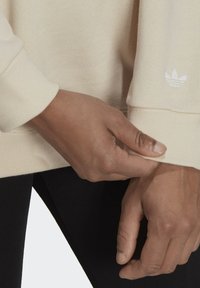 Handen die de polsmanchet van een beige Adidas-trui met het witte trefoil-logo van het merk aanpassen, gedragen met zwarte broek.