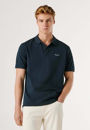 Giovane uomo con capelli corti biondi che indossa una polo blu scuro e pantaloni bianchi, in piedi con una mano in tasca davanti a uno sfondo neutro.