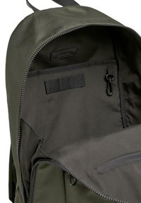 Interno di uno zaino in tessuto verde che mostra una tasca con zip, cinghie in velcro removibili e un moschettone, con una fodera liscia e testurizzata.