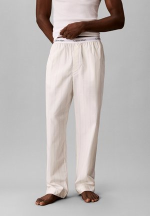 Homme portant un pantalon de pyjama blanc rayé et un débardeur blanc, ajustant la taille montrant le logo Calvin Klein, debout pieds nus sur un sol gris.
