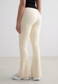 Pantalons légers crémeux évasés avec une texture côtelée, un ourlet festonné et un léger éclat, associés à des baskets grises, montrés de profil.
