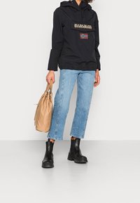 Schwarze wasserdichte Jacke mit einer Fronttasche, versehen mit Logo- und Flaggen-Details; kombiniert mit hellblauen, gerade geschnittenen Jeans und schwarzen Stiefeletten.