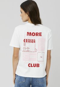 T-shirt en coton blanc avec un design graphique au dos comprenant le texte rouge "PLUS" et "CLUB", ainsi que des illustrations d'un café, d'une tasse de café et d'un croissant.