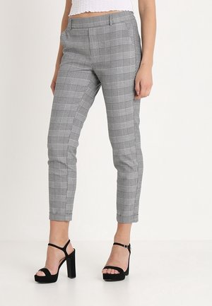 Kvinde iført grå ternede slim-fit ankellange bukser og sorte høje hæle åbne sandaler mod en ensfarvet baggrund.