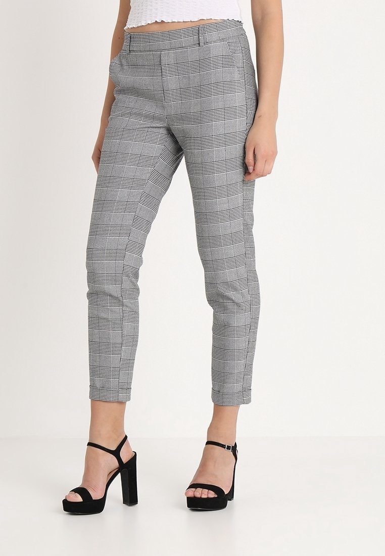 Femme portant un pantalon slim à carreaux gris à la cheville et des sandales noires à talons hauts et bouts ouverts sur un fond uni.
