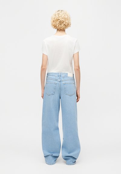 Άτομο με κοντά μπούκλα ξανθά μαλλιά στέκεται με την πλάτη, φορώντας ένα απλό λευκό μπλουζάκι και ανοιχτό μπλε τζιν παντελόνι με φαρδιά μπατζάκια.