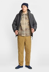Conjunto de hombre que presenta una chaqueta acolchada gris oscura, una camisa beige con estampado, pantalones color arena y un gorro azul marino, junto con zapatillas deportivas azules.