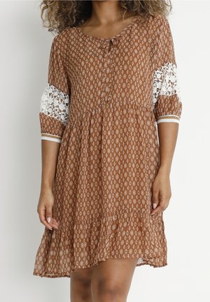 Femme portant une robe marron à motif, longueur genou, avec des manches en dentelle blanche et un lien au niveau du col, debout devant un fond uni.