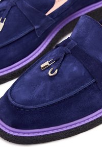 Mocassins en daim bleu marine avec une semelle en caoutchouc violette, ornés d'un nœud décoratif et d'accessoires métalliques pour plus de détails et de texture.