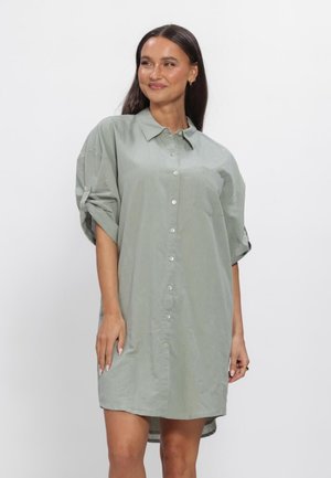 Vrouw met lang donker haar die een losse lichtgrijze blousejurk met opgerolde mouwen draagt, staand tegen een effen witte achtergrond.