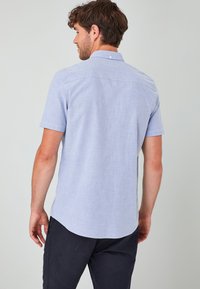 Camisa de botón de manga corta azul claro con una textura suave, dobladillo curvado y detalle en el yugo trasero. Tejido suave con un patrón mínimo.