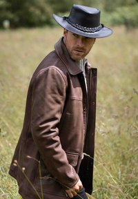 Brown leather jacket with a zip closure and side pockets, paired with a grey sweater, and a black braided leather hat. Grass background.