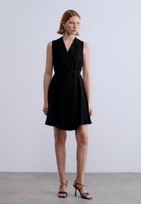 Μαύρο αμάνικο φόρεμα με σχέδιο τύπου κρουαζέ, σφιχτή μέση και φαρδιά φούστα. Φορεμένο με μαύρα πέδιλα με λουράκια. Λείο ύφασμα.